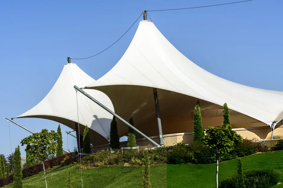 Struktur membrane berpuncak ganda dengan tiang tinggi dan kabel penegang di atas taman berumput