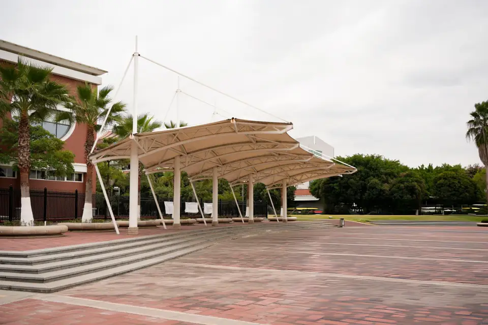 Deretan peneduh membrane melengkung dengan tiang dan kabel penegang di depan gedung sekolah